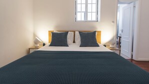 Ein ordentlich bezogenes Bett mit einem karierten Kopfkissen und einer dunkelgrünen Decke, eine Nachttischlampe und ein Fenster mit Jalousien.