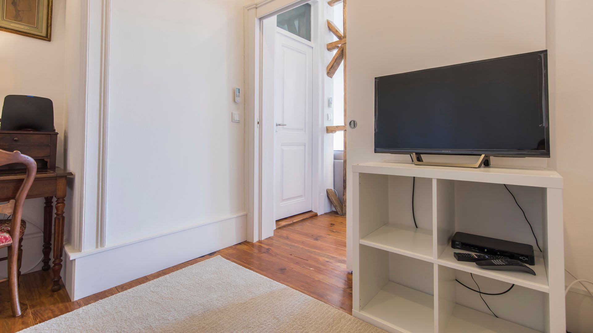 A hallway with a wooden floor, a white door, a television on a white stand, and a small wooden table with a chair.
