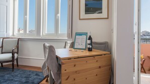 Ein Zimmer mit einem Holztisch, zwei Stühlen, einem gerahmten Bild an der Wand und einem Fenster mit Blick auf Gebäude.