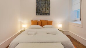 A neatly made bed with white linens and orange pillows, two bedside lamps, and a painting above the bed.