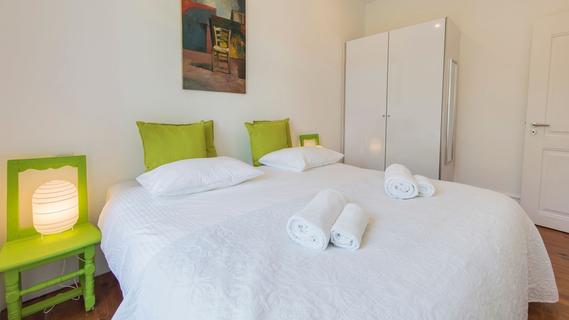 Un lit bien fait, avec du linge de lit blanc et des serviettes pliées, des coussins verts et une table de chevet verte avec une lampe.