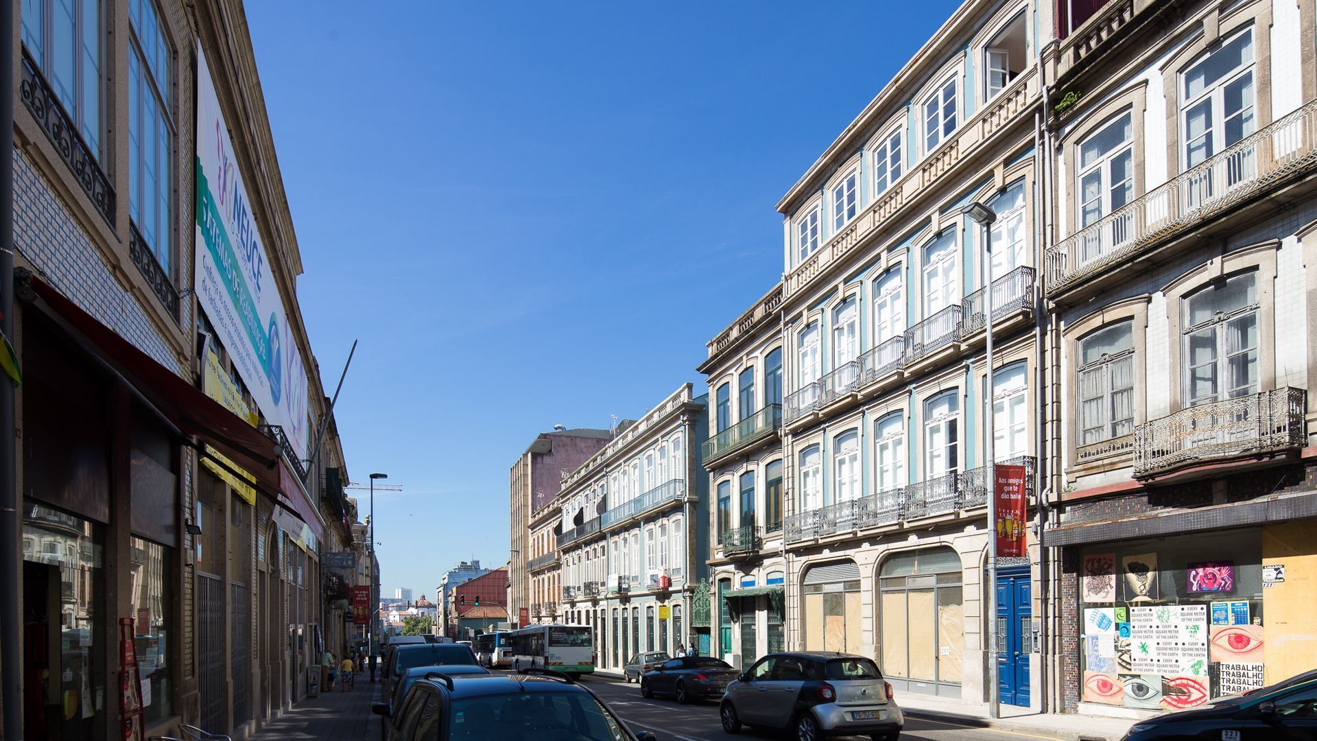 A street lined with multi-story buildings, some with balconies and signage, under a clear blue sky.