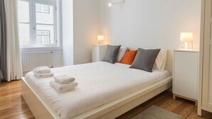 A neatly made bed with white linens and folded towels, a bedside table with a lamp, and a view of a window with curtains. - Downtown Cosy Wonder by LovelyStay (Lisboa)