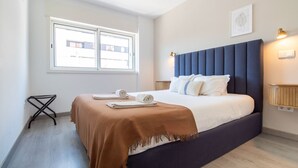 A modern hotel room with a large bed, a window with blinds, a bedside table, and a wall-mounted lamp.