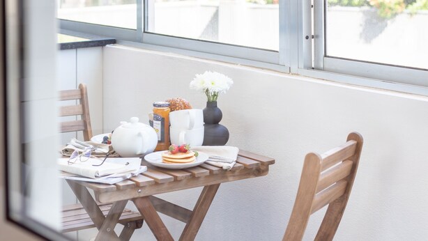 A wooden table with breakfast items, a vase with white flowers, and a window with blinds.