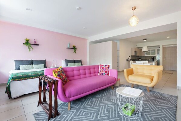 Un salon moderne avec un canapé rose, un fauteuil jaune et un lit recouvert d’une couvre-lit à motifs verts.