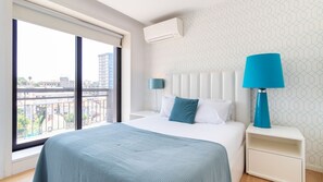 A hotel room with a large window, a bed with white and blue bedding, a blue lamp, and a white nightstand. - Trindade Cozy Apartment by LovelyStay (Porto)