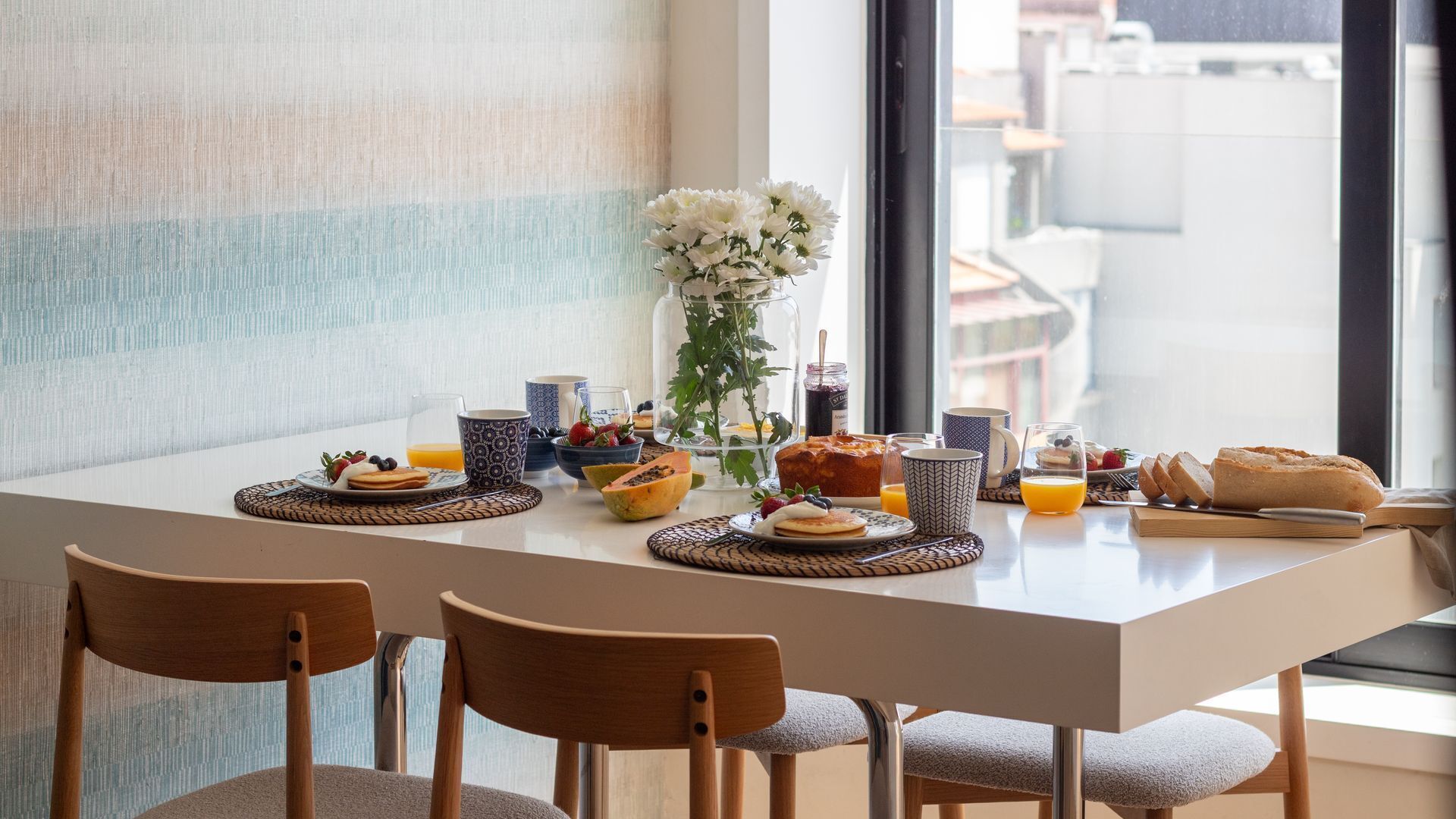 Una mesa de desayuno con diversos alimentos, un florero con flores y una vista de edificios a través de una ventana.