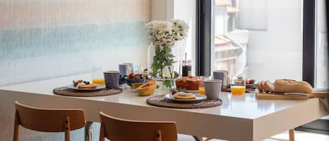 Una mesa de desayuno con diversos alimentos, un florero con flores y una vista de edificios a través de una ventana.