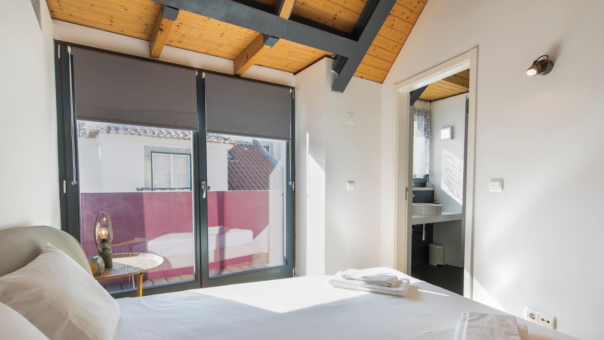 Un dormitorio con un amplio ventanal corredizo que da a un balcón, una cama con ropa de cama blanca y una pequeña mesa redonda con una lámpara.