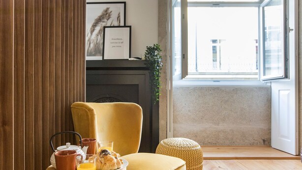 A cozy room with a yellow armchair, a small table with breakfast items, a framed quote on the wall, and a view through a large window. - Mouzinho Studio w/ AC in City Center by LovelyStay (Porto)