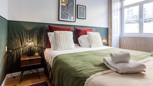 A neatly made bed with a green bedspread, white pillows, and a red pillow. There is a nightstand with a lamp and a small decorative item beside the bed. - Mouzinho Studio w/ AC in City Center by LovelyStay (Porto)