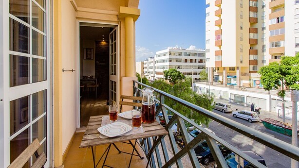 Un balcón con una mesa, dos vasos y una jarra de bebida, con vista a una calle con coches y edificios.