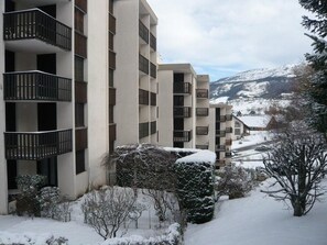 Un immeuble à plusieurs étages doté de balcons, entouré d’arbres couverts de neige et avec une chaîne de montagnes en arrière-plan.