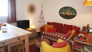 A cozy living room with a wooden dining table, a red sofa, a small round ottoman, and a television on a wooden stand.