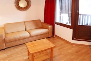 A room with a wooden floor, a beige sofa, a small wooden table, and a window with a view of a balcony.
