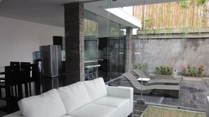 Un área de estar moderna con un sofá blanco, una mesa de centro de vidrio y una cocina visible a través de paredes de cristal.