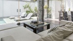 Un salón moderno con un sofá grande, una mesa de centro de vidrio y vistas a una piscina y zona de estar al aire libre.