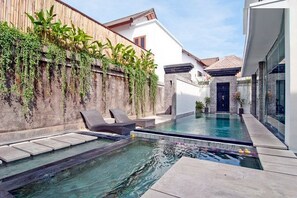 Un área de piscina privada con sillones de descanso, un muro de piedra con follaje y un edificio moderno con tejado de cerámica.