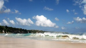 Una playa con olas rompiendo en la orilla, cielo despejado y una costa distante con edificios.