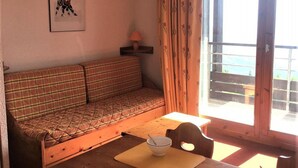A cozy room with a wooden dining table, two bowls, a plaid sofa, and a framed picture on the wall. - 100m from ski slopes, 5th floor, view mountain, swimming pool, balcony, tv, ski locker, 31m² (Le Corbier)