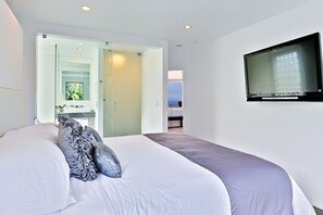 Quarto com cama, televisão fixada na parede e vista para o mar através de uma porta aberta.