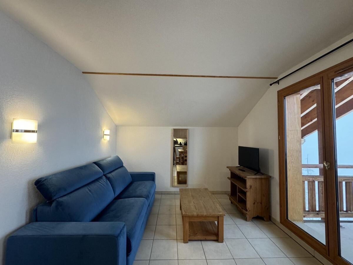 A living room with a blue sofa, a wooden coffee table, and a television on a wooden stand. There is a sliding glass door leading to an outdoor balcony.