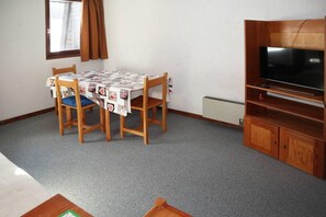 Ein Zimmer mit Esstisch, der mit einer Tischdecke bedeckt ist, Holzstühlen, einem Fernseher auf einem Holzboden und einer Heizung an der Wand.