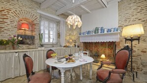Een eetruimte met een witte tafel, twee stoelen, een kroonluchter en een open haard met blauwe theepotten.