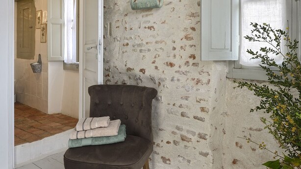 Een rustieke kamer met een houten vloer, een vintage fauteuil met handdoeken erover, een stenen muur en een raam met luiken.