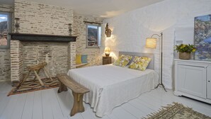 Een slaapkamer met een stenen muur, een bed met bloemenkussens, een bankje, een lamp en uitzicht op het buiten.