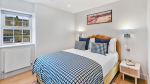 A bedroom with a bed, a window, a radiator, a bedside table, and a wall-mounted lamp. - Cleveland Street by Aeria Apartments (London)