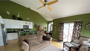 Una sala de estar con un sofá color beige, un sillón marrón y una mesa de centro. Hay una zona de cocina con alacenas blancas y un microondas. La habitación tiene un ventilador de techo y una ventana con cortinas.