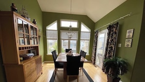 Un comedor con mesa y sillas de madera, un aparador con puertas de vidrio y un amplio ventanal con persianas.