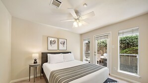 Ein Schlafzimmer mit Bett, Deckenventilator, Nachttisch, Lampe und einem Fenster mit Blick ins Freie.