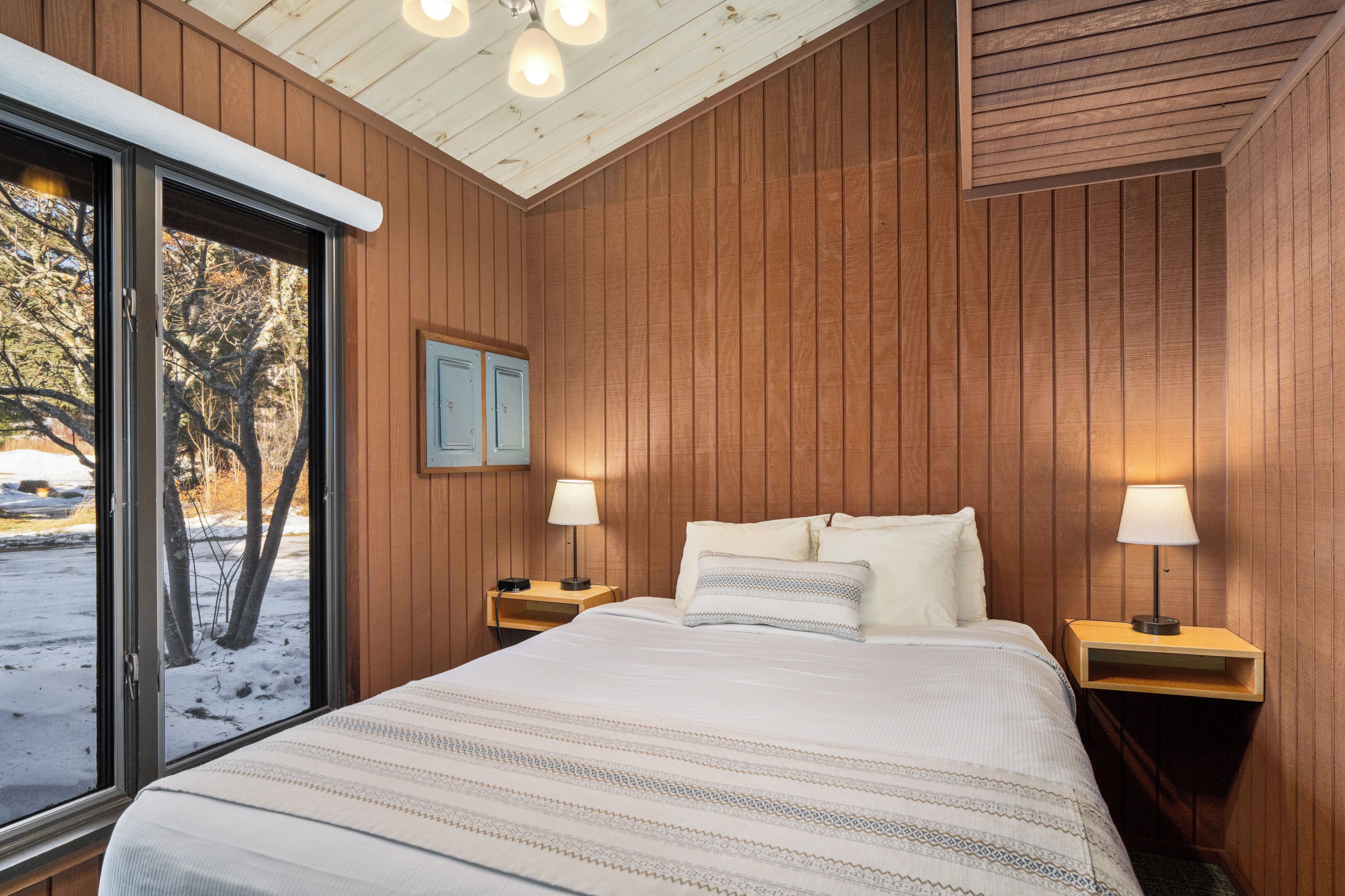 A bedroom with a large bed, wooden walls, a window with a view of trees, and two bedside lamps.