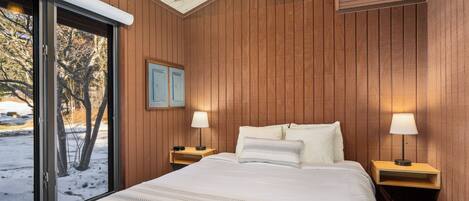 A bedroom with a large bed, wooden walls, a window with a view of trees, and two bedside lamps.