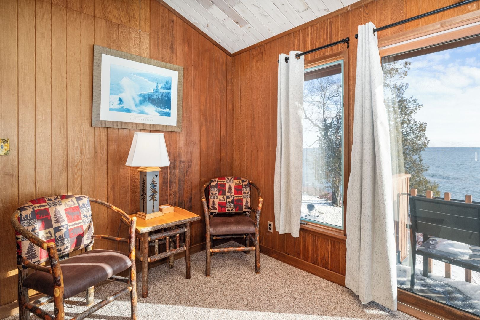 Ein gemütliches Zimmer mit Holzwänden, einem gerahmten Bild, einer Lampe, zwei Stühlen und einem großen Fenster mit Vorhängen.