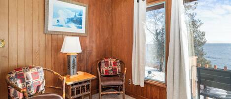 Ein gemütliches Zimmer mit Holzwänden, einem gerahmten Bild, einer Lampe, zwei Stühlen und einem großen Fenster mit Vorhängen.