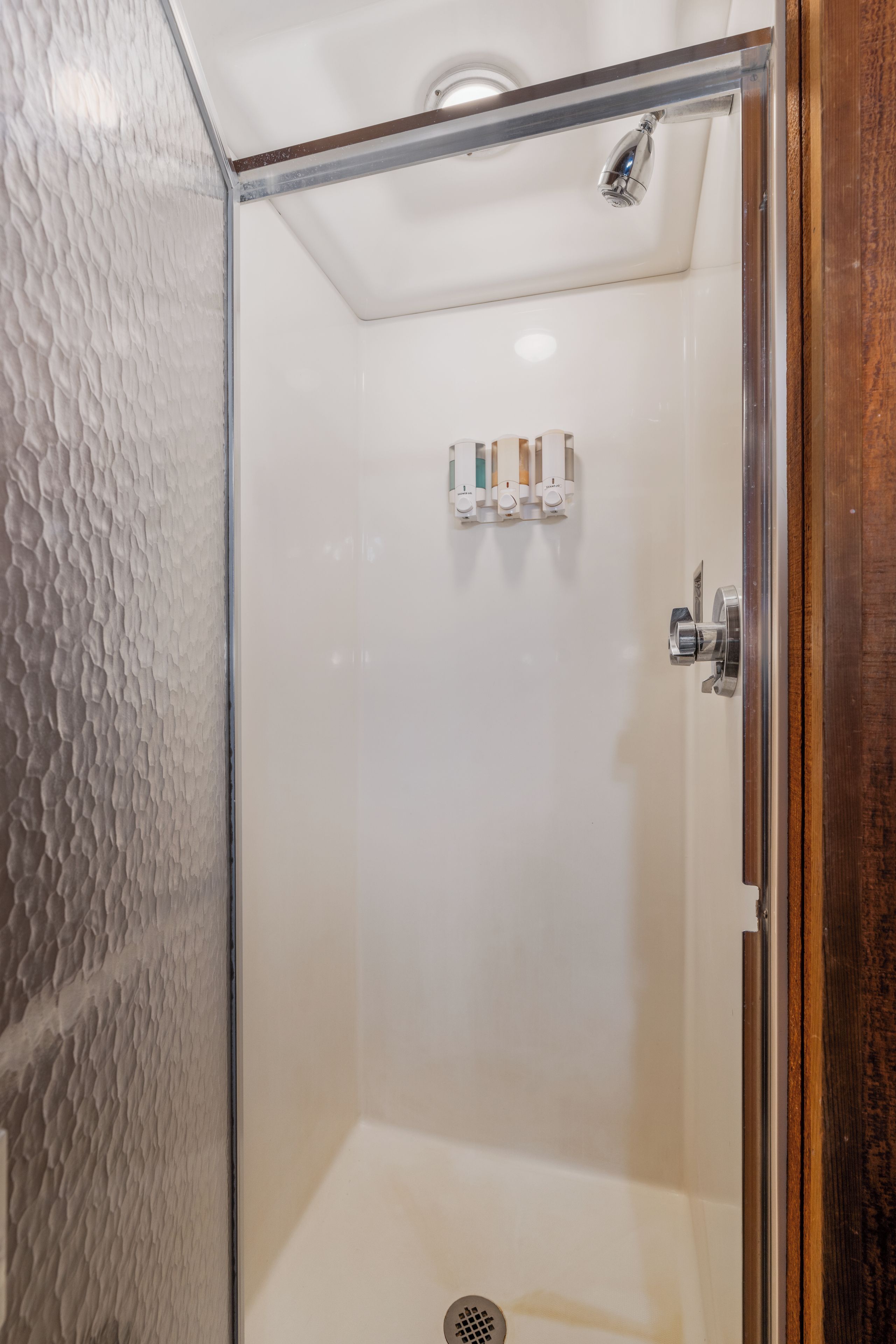 A glass-enclosed shower stall with a white textured wall, a showerhead, and a shelf with toiletries.