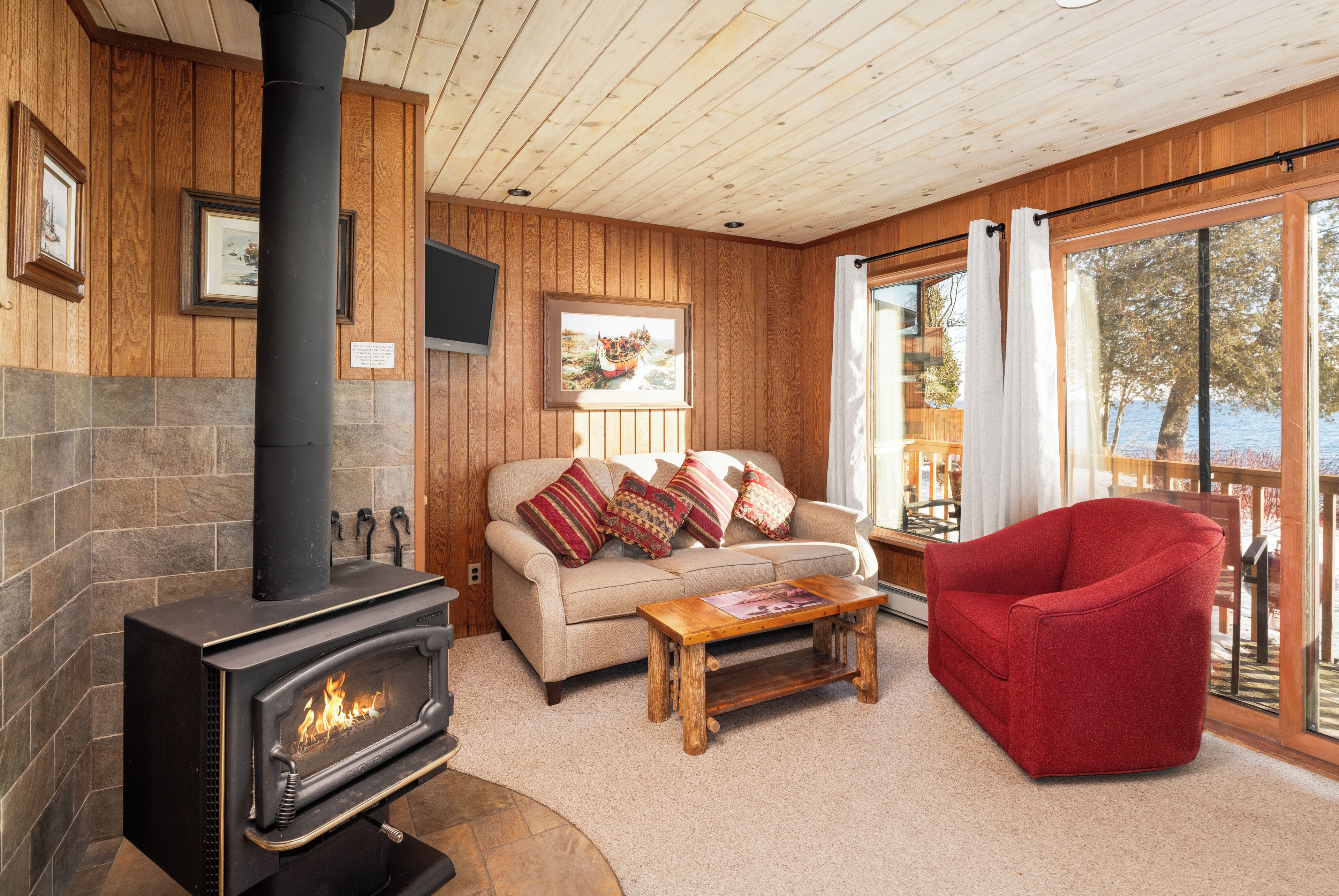 A cozy living room with a fireplace, a sofa with cushions, a red chair, and a large window with curtains.