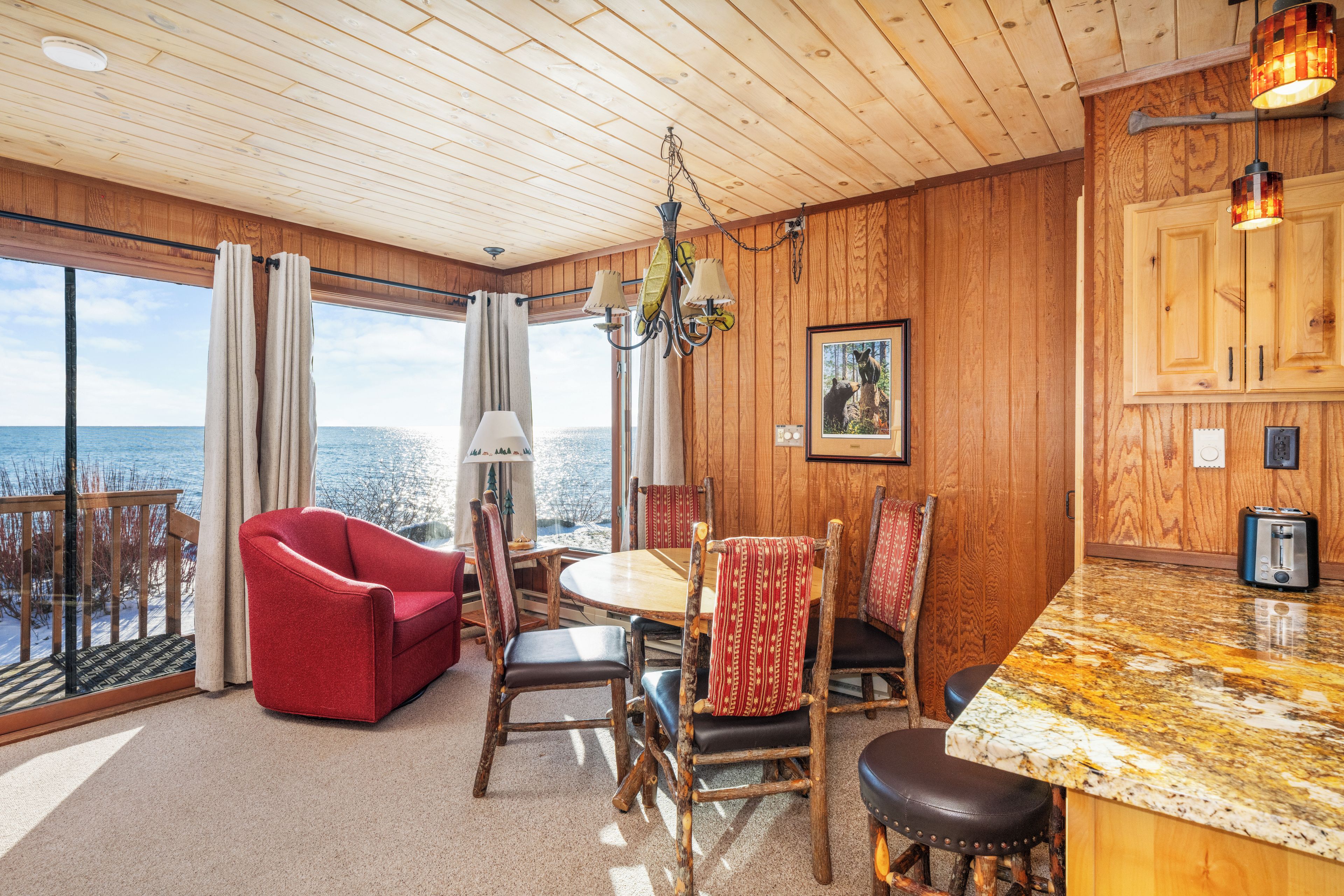 L’intérieur chaleureux d’un chalet avec un plafond en bois, un coin repas comprenant une table et des chaises, une cuisine dotée de meubles en bois et une grande fenêtre offrant une vue sur l’eau.