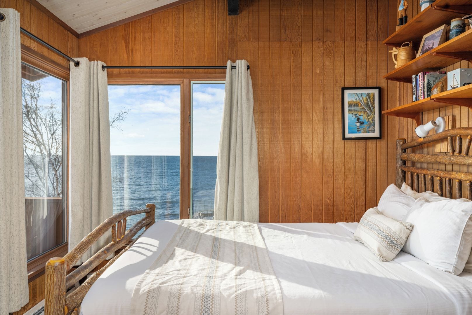 A bedroom with a large bed, wooden walls, a view of the sea, and a bookshelf.