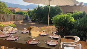 Zona pranzo all'aperto con tavolo imbandito per quattro persone, circondato da sedie, sotto un ombrellone grande.