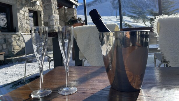 A wooden table with two empty glasses and a metal bucket with a bottle, set outdoors with a stone building and snowy landscape in the background.