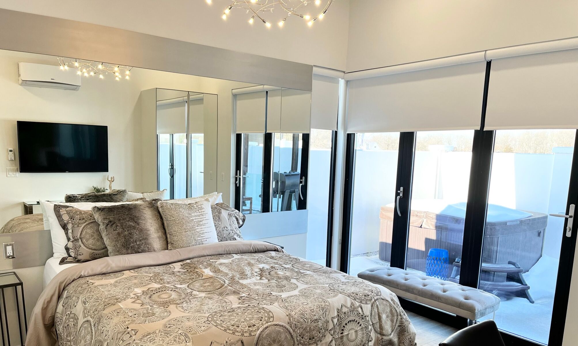 A bedroom with a large bed, a TV, a mirror, and a sliding glass door leading to an outdoor area with a hot tub and barbecue.