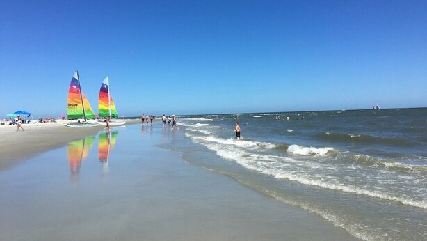 Una playa con veleros, gente caminando y el océano.