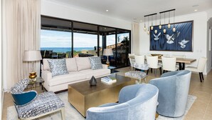 Amplia sala de estar con un gran ventanal con vistas al océano, un comedor con mesa y sillas, y una moderna lámpara de araña.