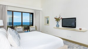 Un cuarto de hotel con una cama grande, un televisor en un estante, vista al océano y una silla junto a la ventana.