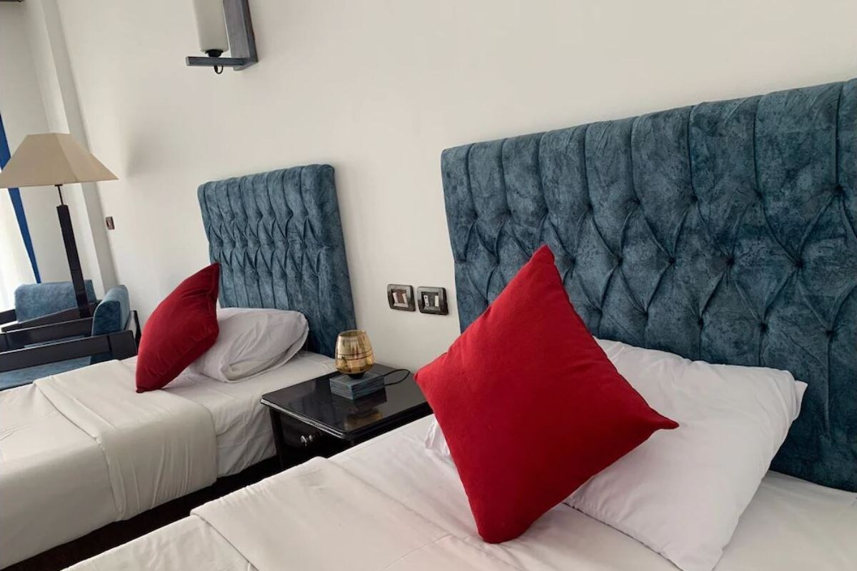Two beds with blue headboards, white bedding, and red pillows.